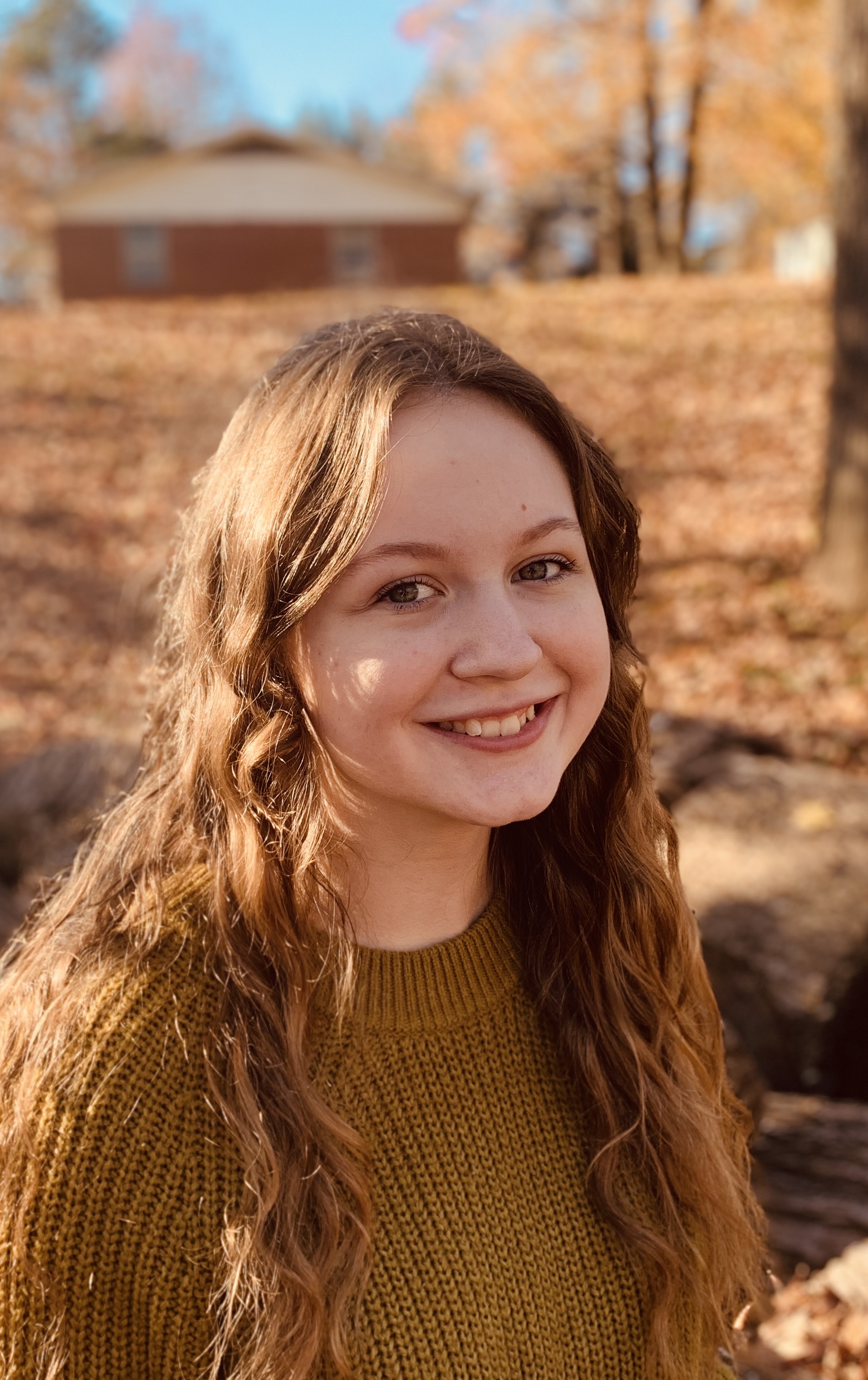 An image of Jorgi Anderson, smiling at the camera. Fall foliage and a red barn stand in the distance.