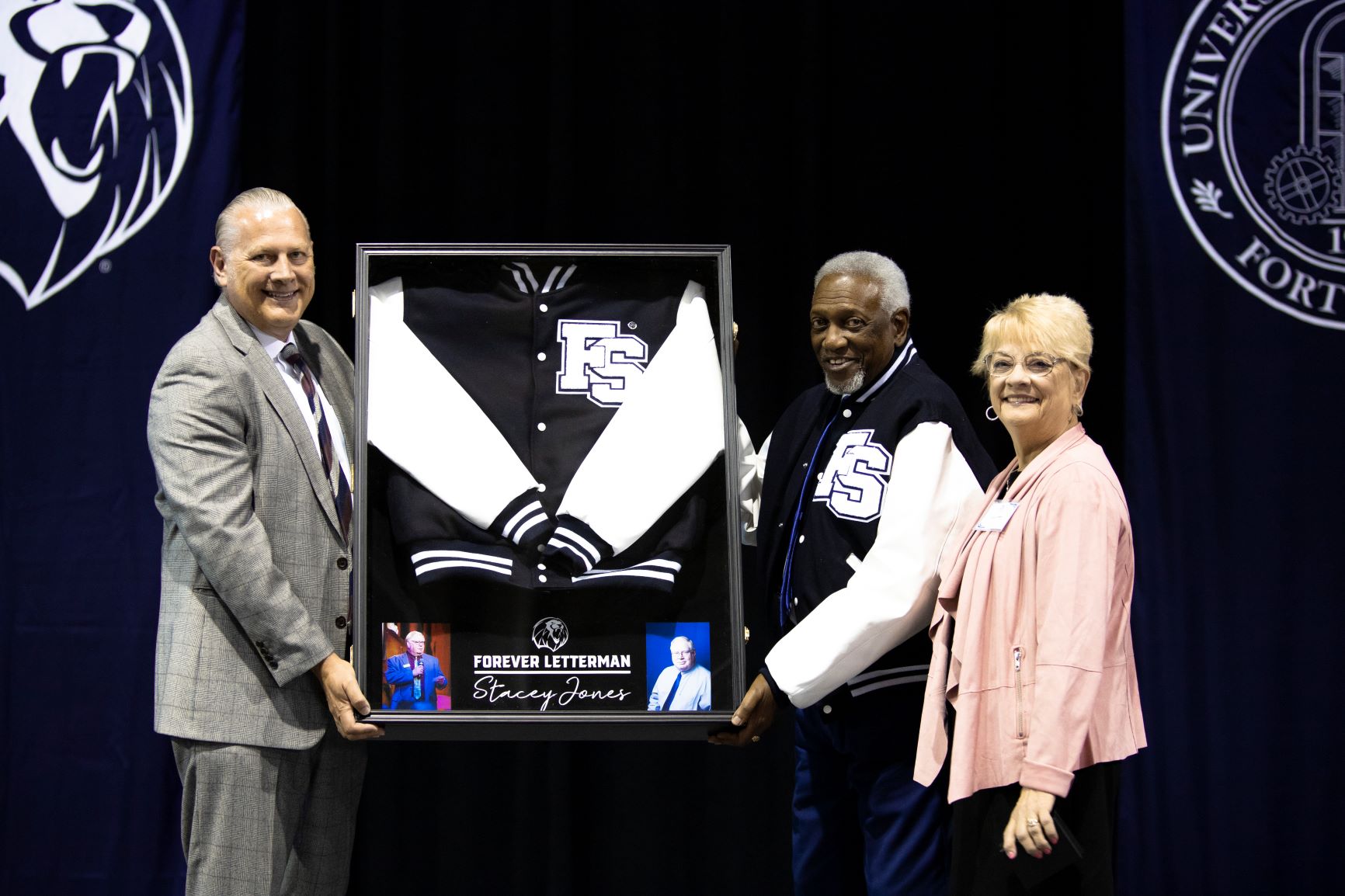 Two men and a woman holding a framed letterman jacket