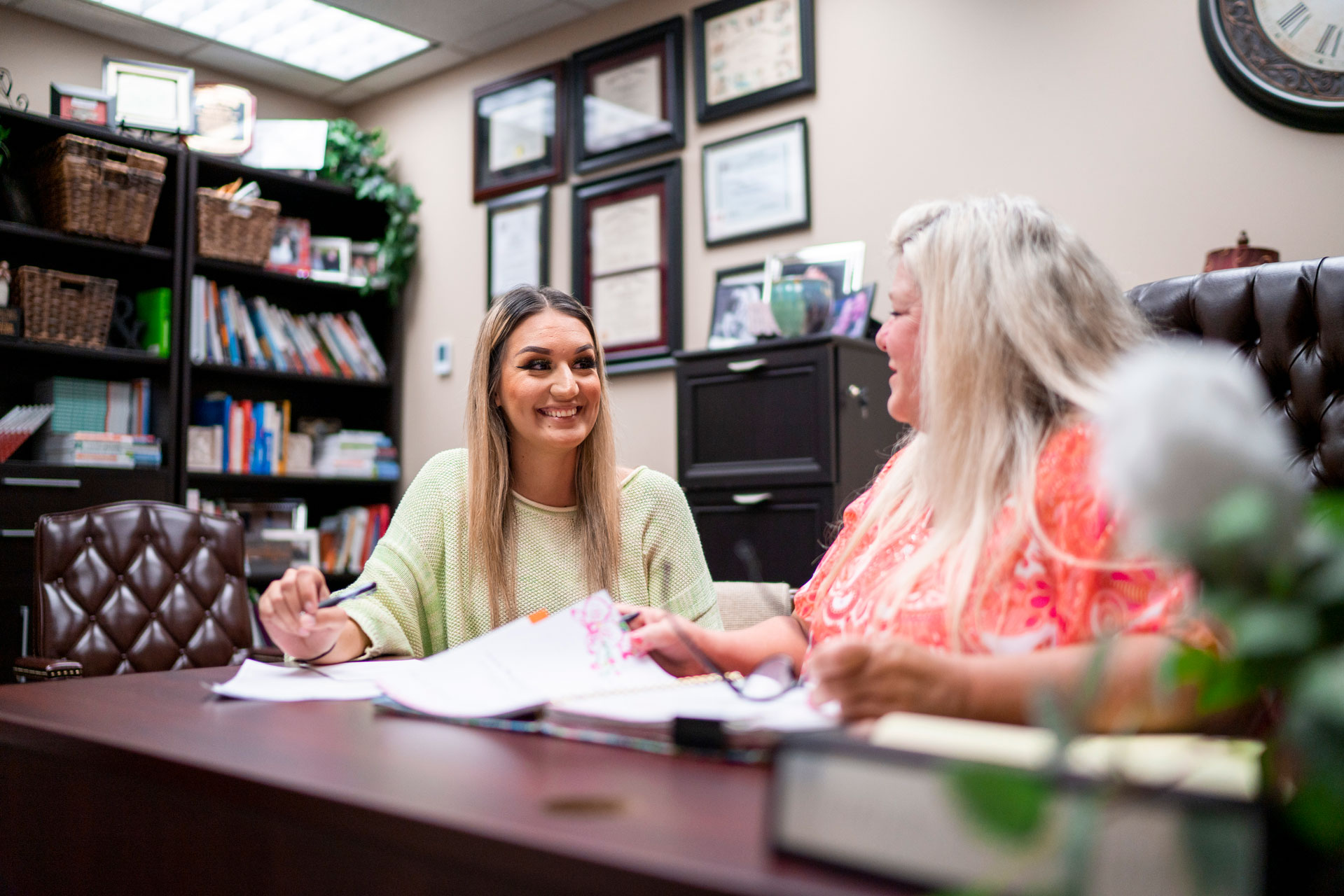 Student sitting at desk smiles as she speaks with another person. 