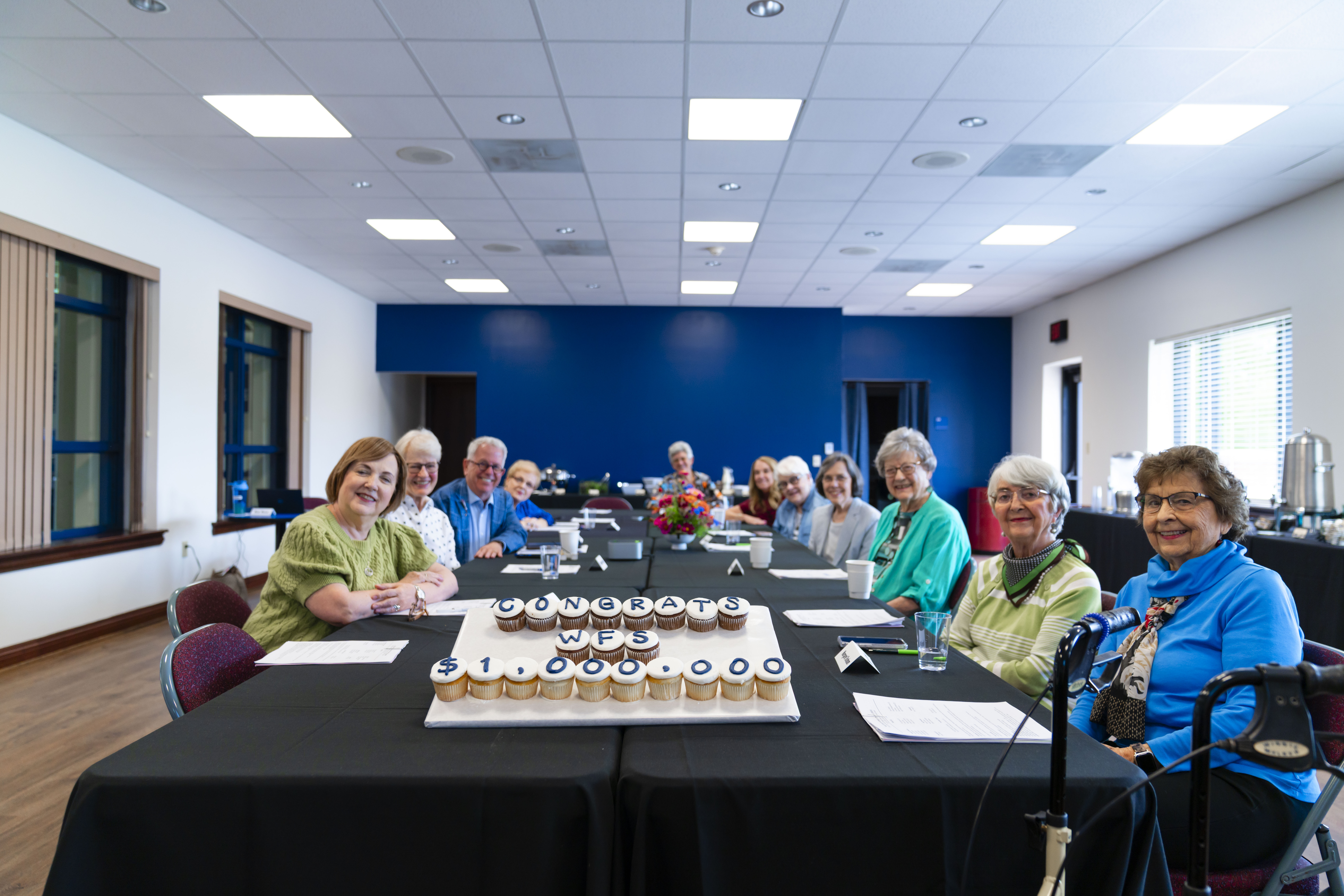 Group of ladies and a man sitting around a table with display of cupcakes.